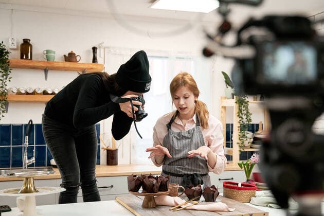 Unrecognizable woman taking photo of chocolate muffins on digital camera against blogger talking during cooking process in kitchen, Model released Copyright: xSergeyxMironovx MR
