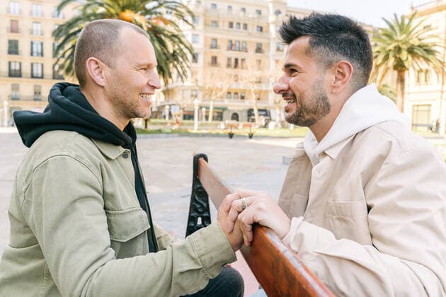 IMAGO Creative: Urban Diversity Side view of joyful young multiracial gay couple resting on wooden bench and looking at each other on city square on sunny day Copyright: xIoritzxLopezx Model Released