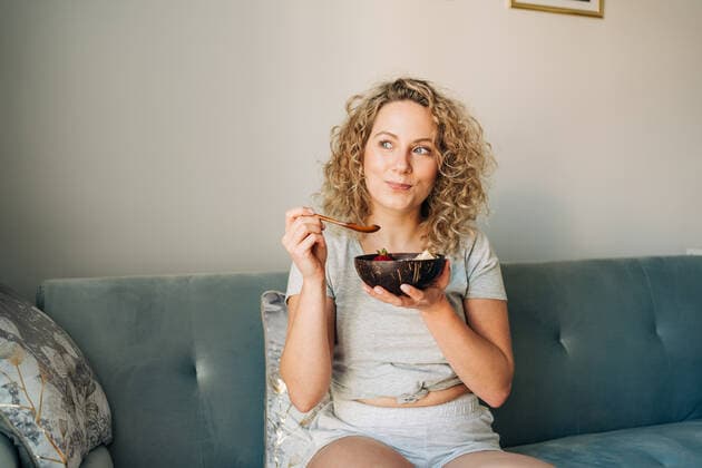 Cheerful female in domestic wear enjoying yummy food in bowl while sitting on comfortable sofa with legs crossed with smile, Model released, Property released Copyright: xJakexJakabx MR