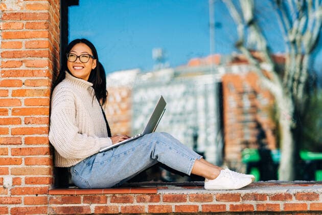 Stylish happy young Asian female remote worker in casual clothes and eyeglasses using laptop while sitting on bench near brick building, Model released Copyright: xJakexJakabx MR