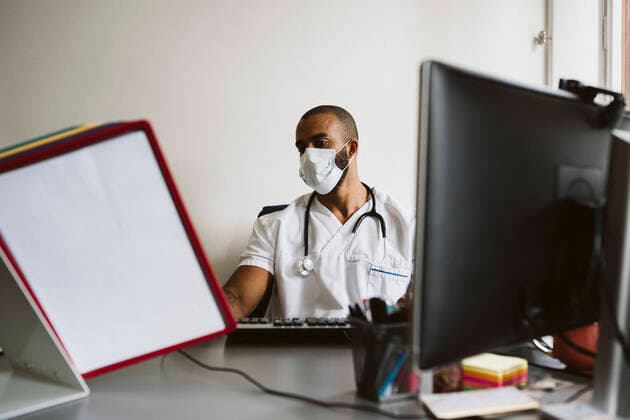 IMAGO Creative: Lokalhelden - Gesundheits- und Rettungskräfte Male doctor wearing protective face mask reading medical files while sitting at desk Sweden, Stockholm, model released, property released Copyright: xMaskotx , MA86797