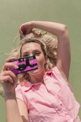 IMAGO Creative, Lifestyle, Barbiecore From above young glad female in pink dress taking pictures on instant photo camera while lying on ground on sunny day Copyright: xAnaxCarpiox Model Released