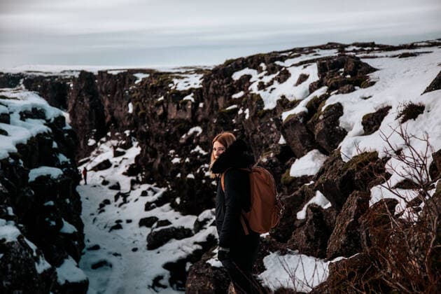 IMAGO Creative: Jahreszeit Winter, Spaziergang Side view of young woman with backpack standing and looking at camera between big snowy rocks in Iceland, Model released Copyright: xEkaitzxGeriquex MR
