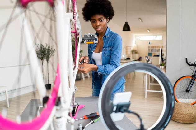 IMAGO Creative: Digitale Welt, Social Commerce Female influencer streaming online on mobile phone while repairing bicycle at home model released Symbolfoto property released GIOF12417