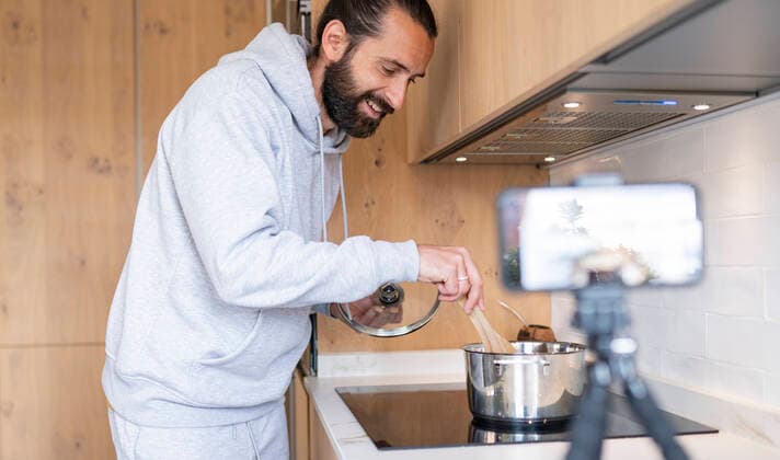 Smiling male influencer vlogging while preparing food in kitchen at home model released Symbolfoto property released JCCMF01938
