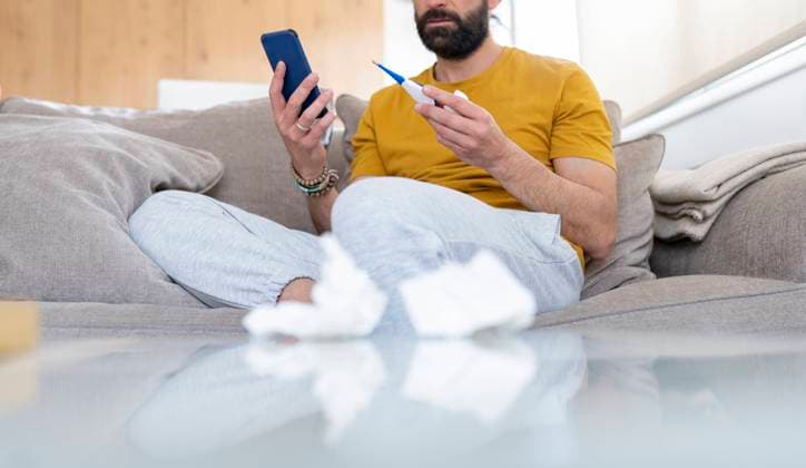 Man holding thermometer and smart phone while sitting on sofa at home model released Symbolfoto property released JCCMF01940