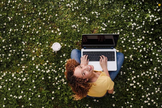 IMAGO Creative: Jahreszeit Frühling, Frühlingsanfang am 20. März IMAGO Creative: Klimawandel - Zukunft und Innovation Creative Highlights Symbolbilder Young woman looking up while sitting with laptop on meadow amidst flowers model released Symbolfoto EBBF03699