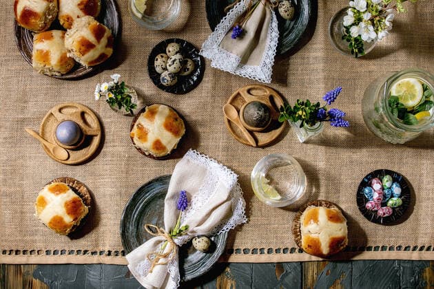 Easter table setting with colored and chocolate eggs, hot cross buns, bouquet flowers, empty ceramic plate with napkin, glass of lemonade drink on wooden table with textile tablecloth. Flat lay NatashaBreen REDA&CO 1028_29_NBR9686