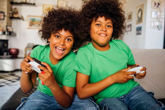 IMAGO Creative, Lifestyle, Gaming Cheerful twin boys with curly hair playing video games sitting on sofa in living room at home model released Symbolfoto property released MEUF03133