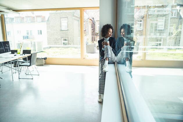 Businesswoman holding coffee cup while sitting on window sill at office model released Symbolfoto property released UUF23526