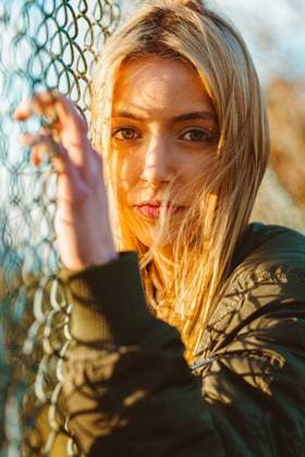 Beautiful blond female in jacket looking at camera standing at chain link fence in golden sunlight, Model released Copyright: xAlbertoxMenendezx MR