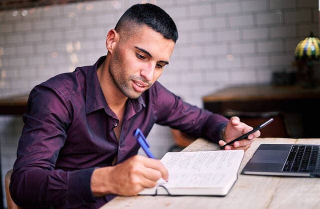 IMAGO Creative: Bildung, Schreiben Side view of handsome young Hispanic male entrepreneur checking information on smartphone and writing notes in planner while working at table with laptop, Model released AdalbertoRodriguez_Businessman_64.jpg Copyright: xAdalbertoxRodriguezx MR