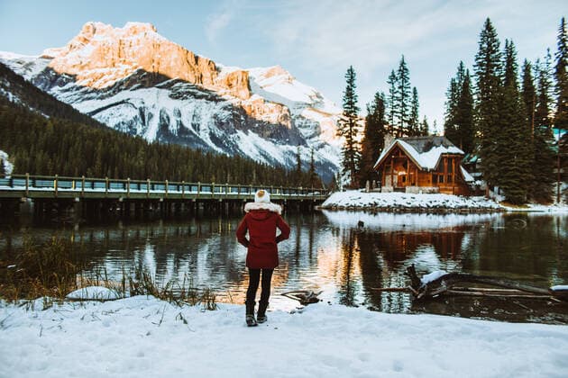 Back view of unrecognizable female tourist in outerwear holding hands in pockets and admiring wooden shack and mountain ridge while walking on snowy coast of Emerald Lake on winter day in British Columbia, Canada, Model released CamilleGirard_Banff_69.jpg Copyright: xCamillexGirardx MR