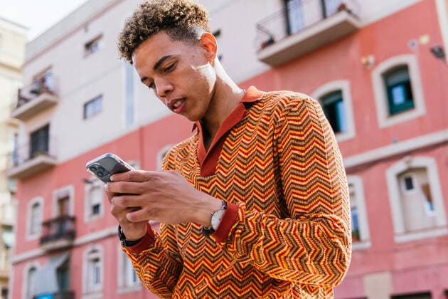 IMAGO Creative: Clickbait, Modetrends From below side view of young Hispanic guy with Afro hair in stylish colorful outfit browsing mobile phone while standing on railing near urban building in sunlight DavidMunoz_JoelBarcelonetaLifestyle_8.jpg Copyright: xDavidxMunozx Model Released