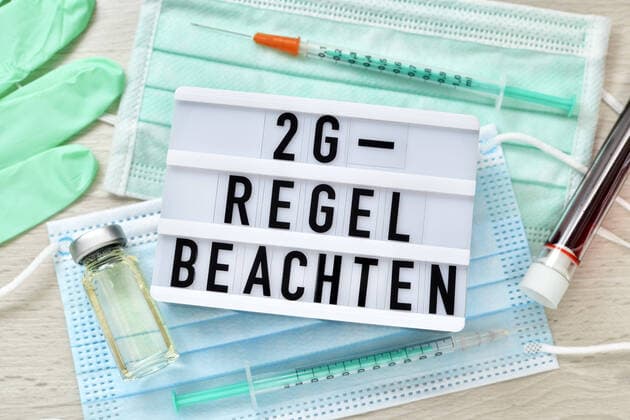 Schild mit Aufschrift 2G-Regel beachten auf Schutzmasken mit Impfstoff und Impfspritzen