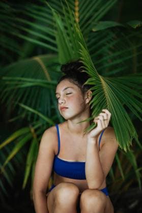 Creative Highlights Symbolbilder Charming young slim female in blue swimwear sitting with eyes closed on tree trunk under green palm foliage and looking at camera while enjoying summer vacation on beach of Costa Rica, Model released CamilleGirard_CostaRicaTravel_63.jpg Copyright: xCamillexGirardx MR