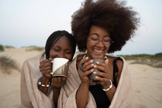 Creative Highlights Symbolbilder Happy young black female best friends with mugs of hot drinks sitting close wrapped in warm blanket and having fun during summer evening on sandy beach OscarDeDios_LifestyleMelDian_29.jpg Copyright: xOscarxDexDiosx Model Released