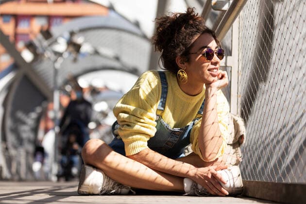 Creative Highlights Symbolbilder Modern millennial Hispanic female with curly hair wearing yellow sweatshirt with denim overalls and trendy sunglasses and earrings sitting leaning on hand looking away near mesh fence in sunlight, Model released SheyNunez_AndreaHomeAndOutdoors_63.jpg Copyright: xSheyxNunezx MR
