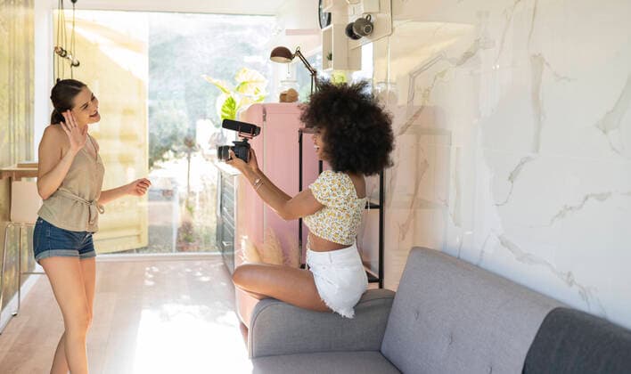 IMAGO Creative: Digitale Welt, Social Commerce Young woman waving in camera held by afro female friend while filming at home model released Symbolfoto property released JCCMF03221