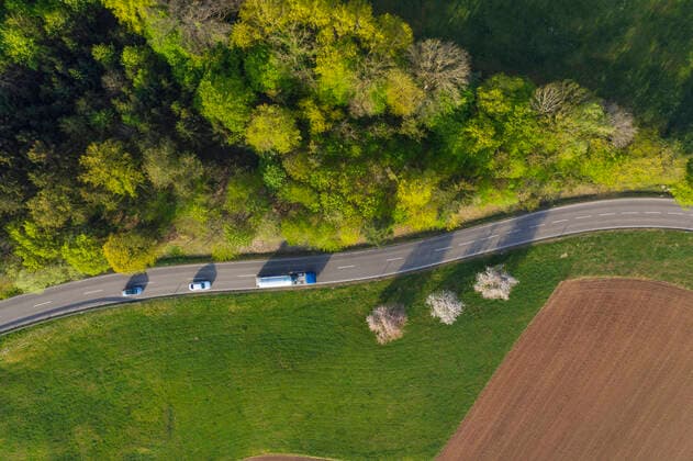 IMAGO Creative: Klimawandel - Zukunft und Innovation Drone view of cars driving along countryside road stretching between green grove and plowed field RUEF03355