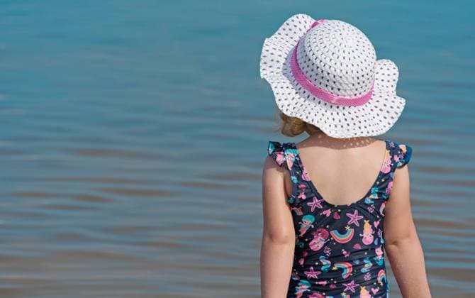 Junges Mädchen steht am Strand und blickt aufs Wasser Kind mit Sonnenhut im Sommer am Badesee. *** Young girl standing on the beach looking at the water child with sun hat in summer at swimming lake