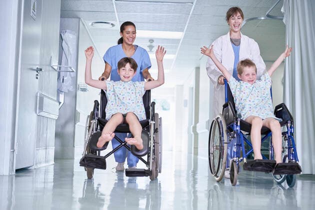 Medical orderlies having fun pushing boy patients in wheelchair on hospital children s ward, 05.07.2016, Copyright: xFrankxandxHelenax, model released, wearing,pushing,playing,working,front view,full length,mischief,childhood,responsibility,bonding,care,cooperation,fun,innocence,skill,teamwork,trust,on the go,25 to 29 years,child,caucasian ethnicity,mixed race ethnicity,multi-ethnic group,woman,young woman,boy,colleague,four people,medical occupation,nurse,patients,indoors,hospital,examination gown,lab coat,surgical scrubs,arms raised,illness,medical,pediatrics,fooling around,transport,wheelchair,6 to 7 years,health service,patient,enjoyment,both male and female,Star Images,healthcare,medical orderly,9 to 10 years,July 2016 Release,chil Ref:IS09AZ9GD