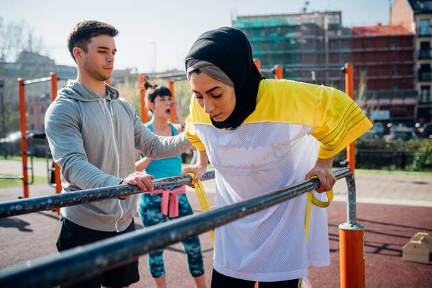 IMAGO Creative: Sport in der Stadt Calisthenics class at outdoor gym, male trainer encouraging young woman on parallel bars, 15.05.2019, Copyright: xEugenioxMarongiux, model released, wearing,exercising,exercise class,push-ups,recreation,using,front view,waist up,concentration,determination,real people,athleticism,bonding,encouragement,fitness,guidance,healthy lifestyle,sharing,support,teamwork,togetherness,well being,friendship,on the go,exercise equipment,parallel bars,20 to 24 years,25 to 29 years,caucasian ethnicity,mixed race ethnicity,multi-ethnic group,female,woman,young woman,male,man,young man,three people,personal trainer,italy,milan,outdoors,park,urban,day,hooded top,t-shirt,hijab,sportswear,sport,active lifestyle,building exterior,both male and female,public h Ref:IS09C0J42