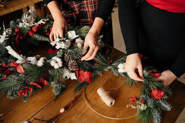 Copped unrecognizable female friends standing at wooden table making creative Christmas wreath from decorative fir and red ribbons for holiday celebration, Model released, Property released GustavoPozo_ChristmasDecoration_40.jpg Copyright: xGustavoxPozox MR