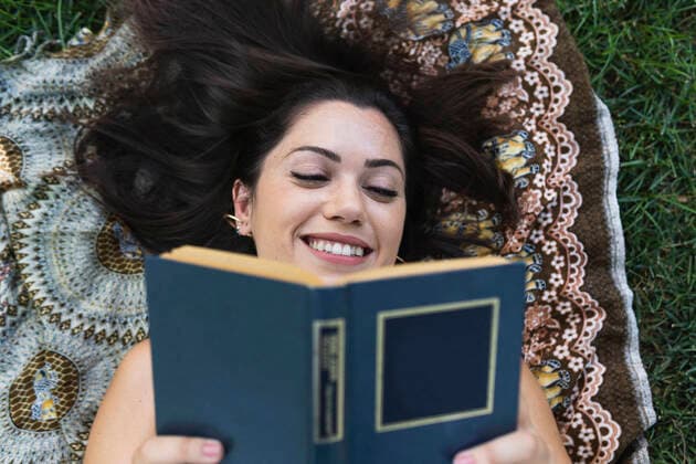 IMAGO Creative: Kulturelle Folgen der Pandemie: Gesundheitsbewußtsein und Alleinsein Smiling young woman reading book while lying on blanket at park model released Symbolfoto PNAF02189