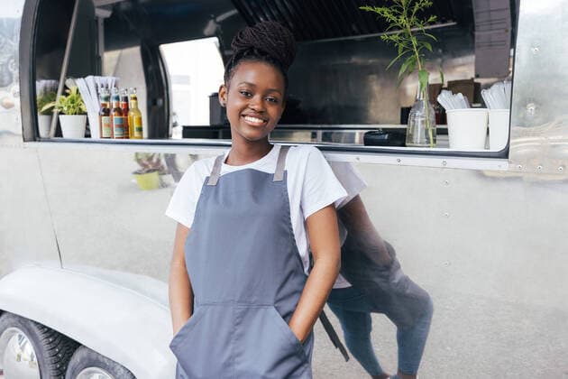 Portrait of smiling female owner in apron leaning on a food truck and looking at camera Copyright: xxArtemxVarnitsinx