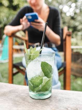 IMAGO Creative: Sommer, Sommertage im Garten Glass of refreshing cocktail with ice and mint leaves placed on wooden table on blurred background of woman browsing smartphone in summer garden, Model released AlbertMorcillo_Finalsummer_19.jpg Copyright: xAlbertxMorcillox MR