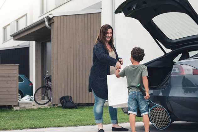 IMAGO Creative: Internationaler Frauentag, Mental Load und Care-Arbeit Smiling mother giving bag to son standing by electric car at front yard Sweden, Skane, model released, property released Copyright: xMaskotx , MA89552