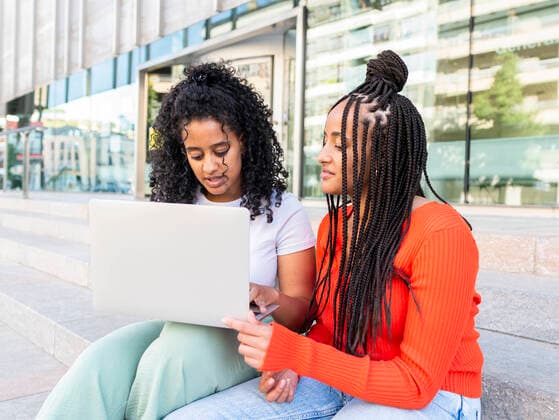 IMAGO Creative: Urban Diversity Cheerful diverse female friends in trendy clothes sitting on stair and browsing laptop on city street in summer day, Model released AlbertMorcillo_MultiethnicFriends_16.jpg Copyright: xAlbertxMorcillox MR