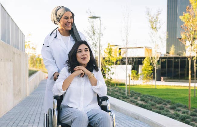 IMAGO Creative, Diversity, Pflege im Krankenhaus Cheerful female healthcare worker standing behind disabled woman sitting on wheelchair model released, JCCMF04282