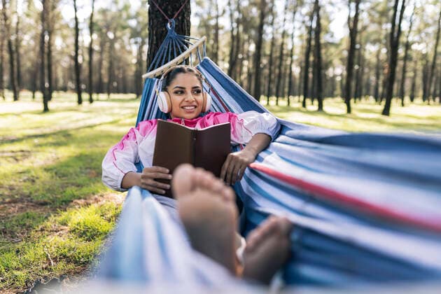 Smiling woman with book lying in hammock listening music at park model released Symbolfoto JCCMF04577