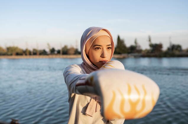 IMAGO Creative: Sport in der Stadt Serious Arab female in traditional hijab and boxing gloves looking away while practicing punching on embankment near rippling river JoseCarlosCerdeno_ArabWomanSportPark_27.jpg Copyright: xJosexCarlosxCerdenox Model Released