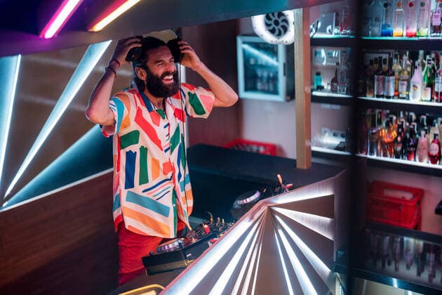 Creative Highlights Symbolbilder Cheerful young bearded male DJ in colorful shirt and headphones laughing while mixing music at counter in contemporary illuminated nightclub JavierDeLaTorre_DJTeatre_41.jpg Copyright: xJavierxDexLaxTorrex Model Released