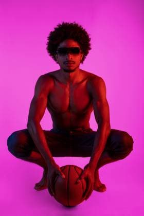 Creative Highlights Symbolbilder Full body of young African American male with curly hair and sunglasses squatting against pink background while holding basketball, Model released SheyNunez_MichelBasket_9.jpg Copyright: xSheyxNunezx MR