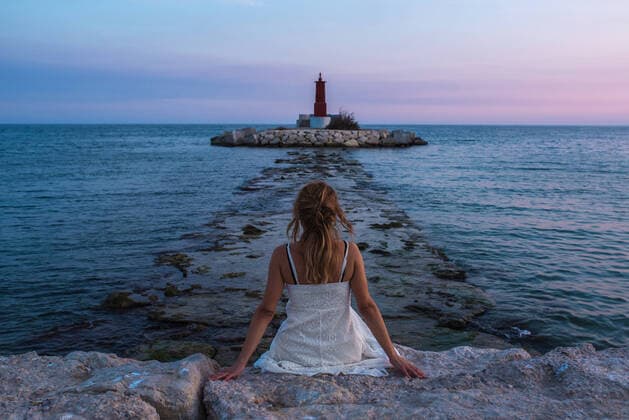IMAGO Creative: Welttourismustag am 27. September Back view of unrecognizable female traveler in sundress sitting on rocky surface against lighthouse in rippling sea during vacation in Spain FranciscoCarrodeguas_WomanAtAlicante_21.jpg Copyright: xFranciscoxCarrodeguasx Model Released