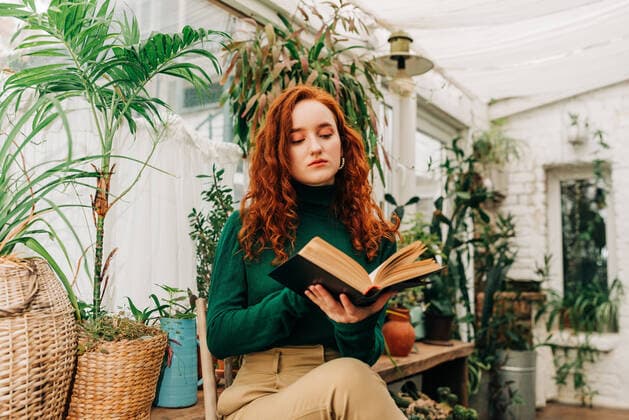 IMAGO Creative: Kulturelle Folgen der Pandemie: Gesundheitsbewu?tsein und Alleinsein From below of concentrated red haired female sitting on chair with crossed legs and reading book among different potted plants in domestic garden OlgaKrivokoneva_WomanInEcoHome_15.jpg Copyright: xOlgaxKrivokonevax Model Released