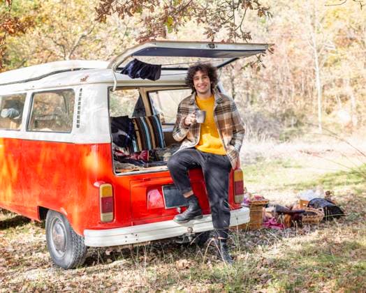 IMAGO Creative: Herbst, Touren mit dem Camper Smiling young man with coffee mug sitting in van s trunk model released, EIF02412