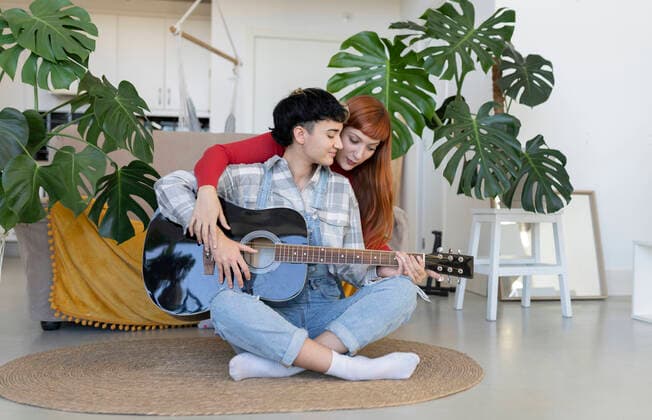IMAGO Creative: Bildung, Musikerziehung Content lesbian hugging talented female partner playing on acoustic guitar on rug in light living room with green potted plants JoseCarlosIchiro_CoupleWomaenAtLoft_37.jpg Copyright: xJosexCarlosxIchirox Model Released