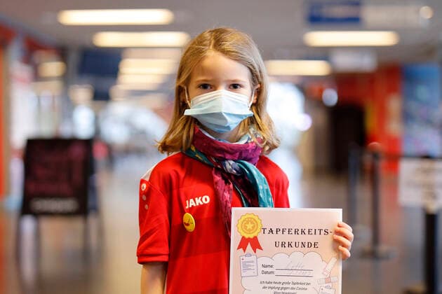 Ein vollständig geimpftes Kind mit Tapferkeitsurkunde bei der Vorstellung der Buttons von Thomas Baumgärtel mit der Impfbanane im Impfzentrum der Stadt Köln in der Lanxess-Arena. Hier werden auch Kinder (5-11 Jahre) geimpft. Vollständig geimpfte Kinder erhalten als Geschenk einen Button sowie eine Tapferkeitsurkunde. Die Buttons mit der Impfbanane sollen zudem an zahlreichen weiteren Impfstellen verteilt werden. Köln, 07.01.2022