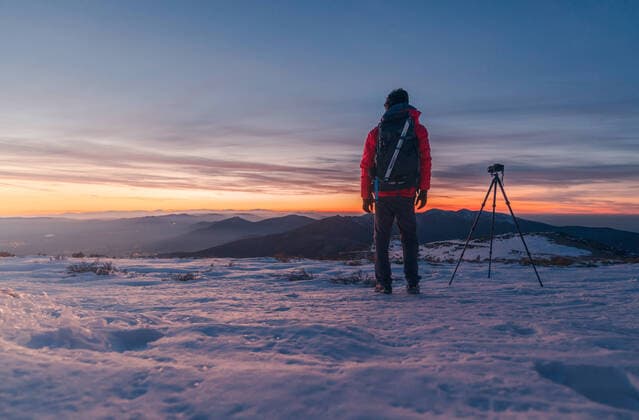 IMAGO Creative: Winter spazieren Full body back view of anonymous traveler standing on snowy ground near tripod with camera while admiring mountains on cold winter evening, Model released JavierMartinez_BolaDelMundo_24.jpg Copyright: xJavierxMartinezx MR