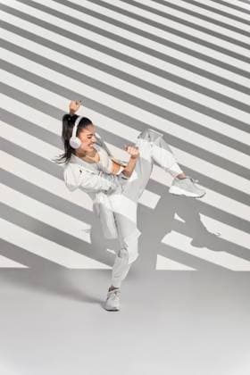 Full body of female dancer in sportswear performing freestyle dance with wireless headphones while enjoying music in studio with stripes of shadows during rehearsal with leg raised, Model released ManuelRuiz_DancerWhiteStudio_22.jpg Copyright: xManuelxRuizx MR