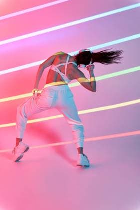 Back view of unrecognizable female dancer in sportswear performing freestyle dance with wireless headphones while enjoying music in studio with neon stripes during rehearsal, Model released ManuelRuiz_DancerWhiteStudio_30.jpg Copyright: xManuelxRuizx MR