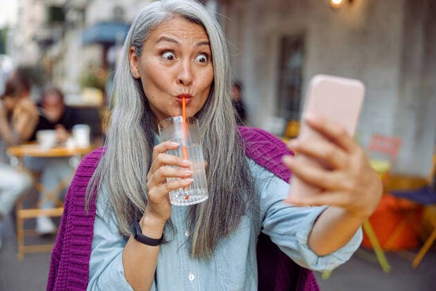 IMAGO Creative: Internationaler Frauenmonat, Gesundheit, Frauen in der Menopause Funny mature Asian woman drinks water taking selfie with cellphone in cafe Funny mature Asian woman with long hair drinks water through straw taking selfie with modern cellphone on outdoors cafe terrace Copyright: xyaroslavxastakhovx 40965918