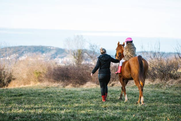 IMAGO Creative, Kinder auf dem Reiterhof Full body back view of anonymous female helping faceless kid in helmet riding brown horse on grassy lawn in countryside, Model released HodeiUnzueta_WinterHorses_44.jpg Copyright: xHodeixUnzuetax MR