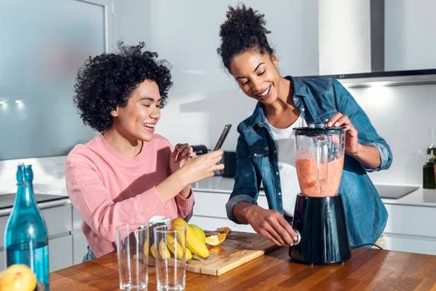 Smiling woman photographing friend blending juice in kitchen at home model released property released, JSRF01909