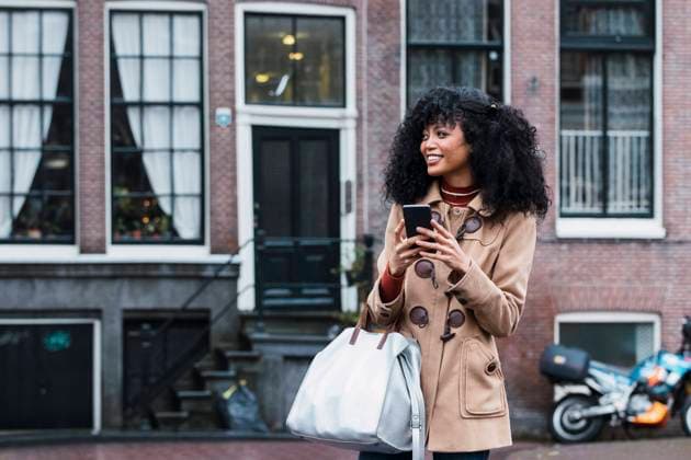 IMAGO Creative: Welttourismustag am 27. September Happy young woman with purse holding smart phone in city model released, PNAF03357